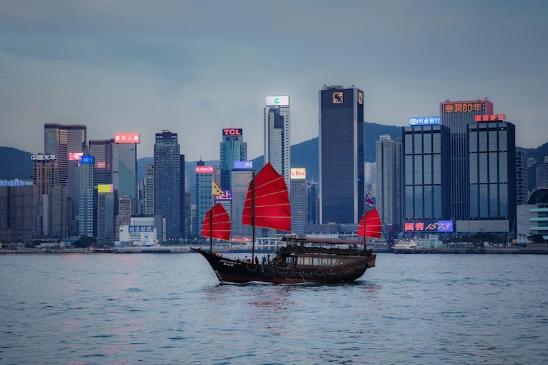 A traditional junk boat with red sails floats in front of the Hong Kong city skyline.
