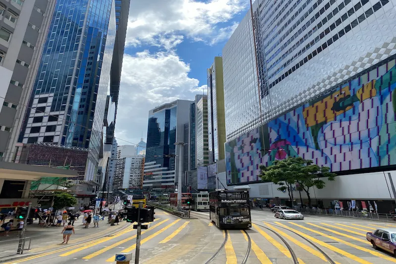City street with tall glass buildings, a double-decker bus, yellow road markings, and pedestrians under a cloudy sky.