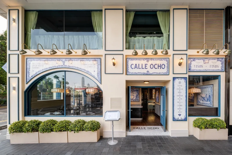Calle Ocho restaurant exterior in Hong Kong with large windows, green shrubs, and blue sign above the entrance.