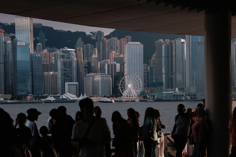 People gather by the waterfront with the Hong Kong skyline and Ferris wheel illuminated at dusk.