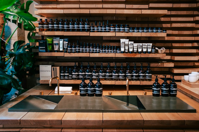 Wood shelves with skincare products in a 2025 retail store, featuring gold faucets and green plants.
