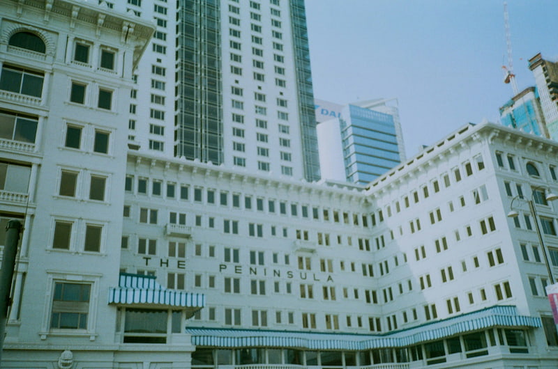 白色建築物掛有The Peninsula招牌及藍色間條遮篷，背景為香港摩天大廈。