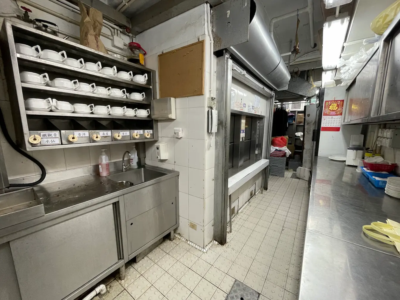灣仔商用廚房設金屬工作枱、放滿杯子的層架、洗手盆、地磚及整齊擺放的廚具。