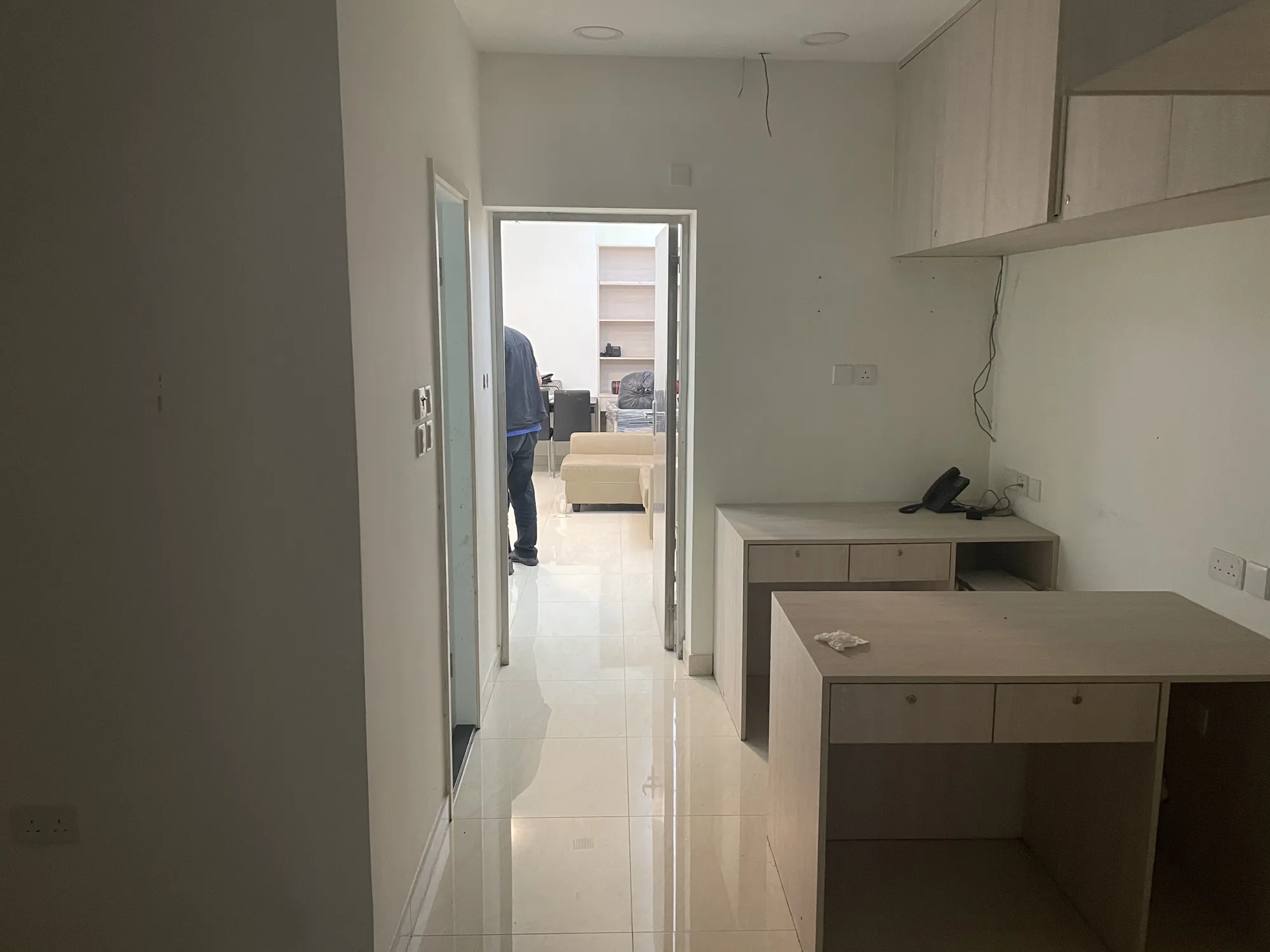 A hallway with built-in desks leads to a central room with two people by a beige sofa and wall shelves.