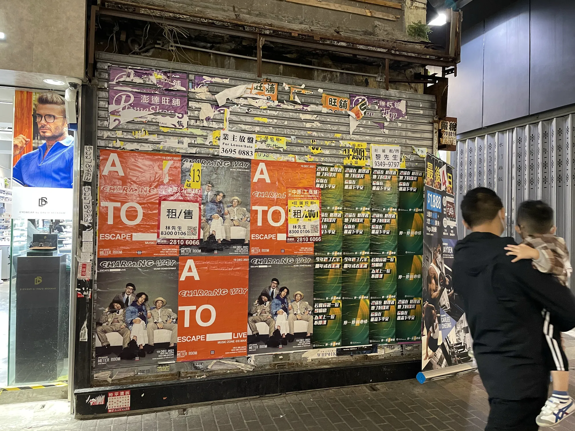 Posters and flyers in Chinese cover the metal shutter of a closed storefront at night, hinting at a rental opportunity. Two people pass by on the right, one carrying a child. Bright shop lights illuminate Ref#3159.