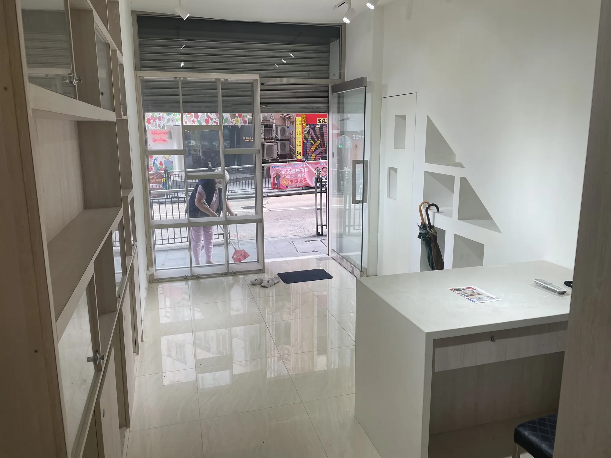 Small empty shop off Hollywood Road with tiled floor, a counter, and a person sweeping by the open Central entrance.
