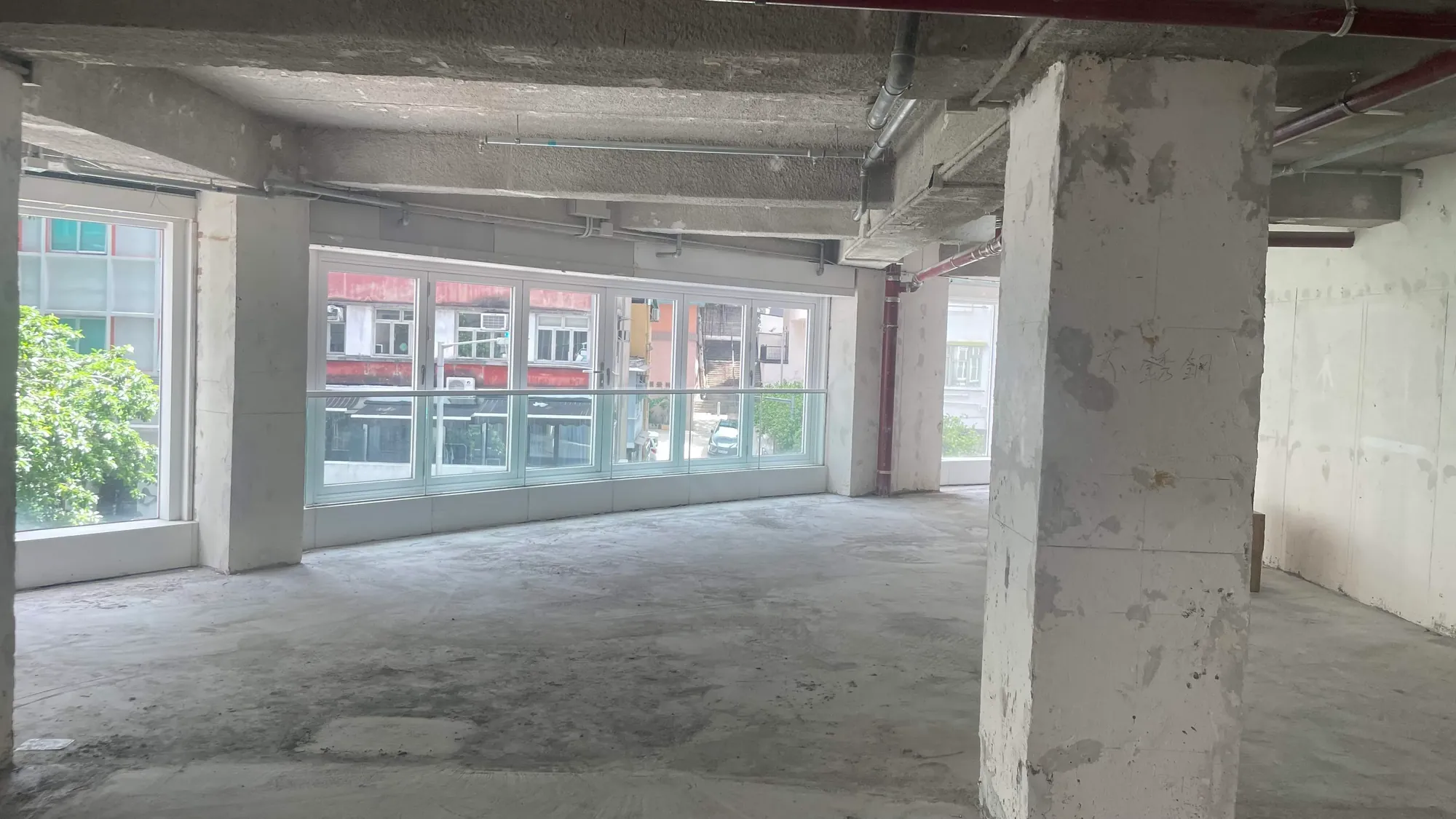 Ref#2990: Spacious, unfinished room with large windows for natural light ideal to rent as office or shop space. Features concrete floor, exposed ceiling, structural columns; buildings and greenery visible outside.