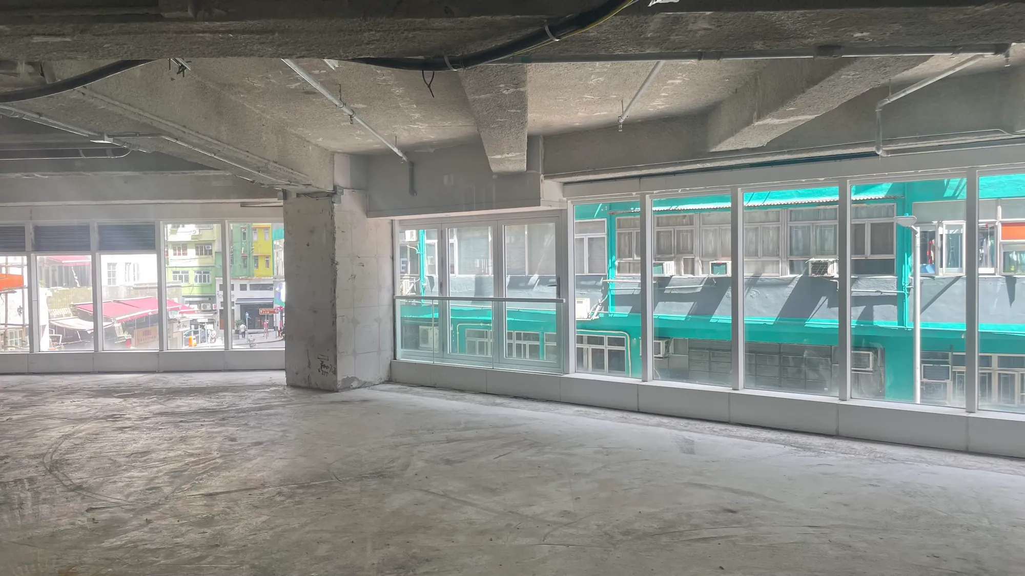 Ref#2990 features a spacious, empty room with unfinished concrete floors and ceiling, large windows for natural light, and views of colorful buildings on a lively street ideal to rent as a shop or office.