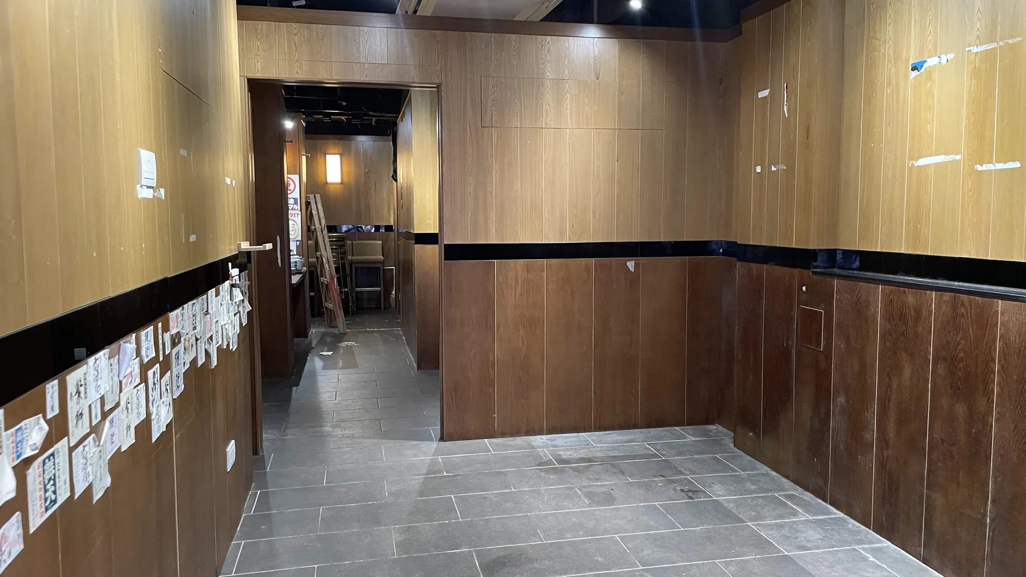 Ref#2995 features a mostly empty hallway with wood-paneled walls, tiled floor, and notes on the left wallgreat for office or shop rental. Overhead lights brighten the space, with an open doorway to a dimly lit area with chairs and tables in back.
