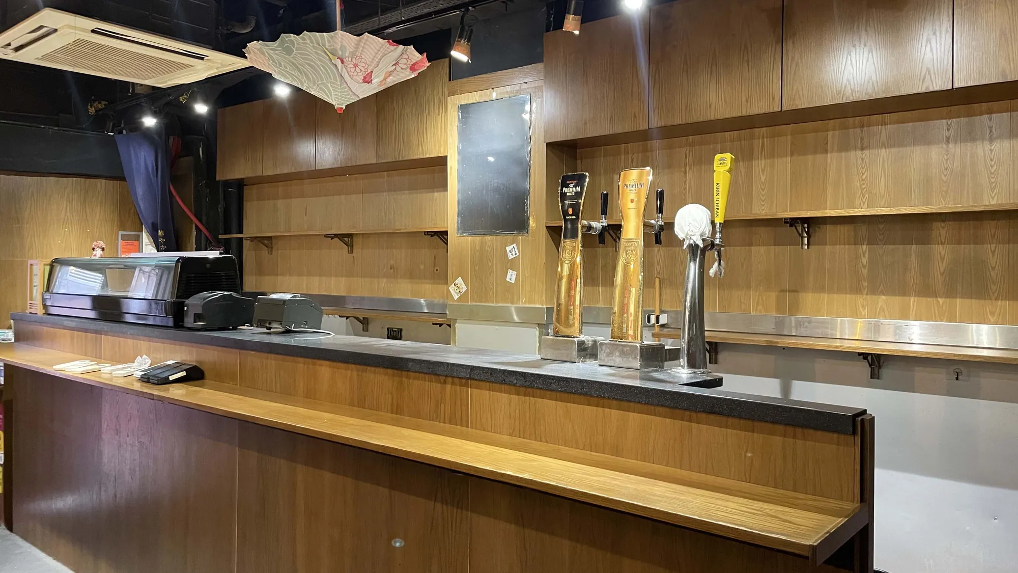 Ref#2995: Wooden bar counter with empty shelves and beer taps, no people present. The dimly lit bar appears closed or being prepped for restaurant rental, with an umbrella hanging from the ceiling.
