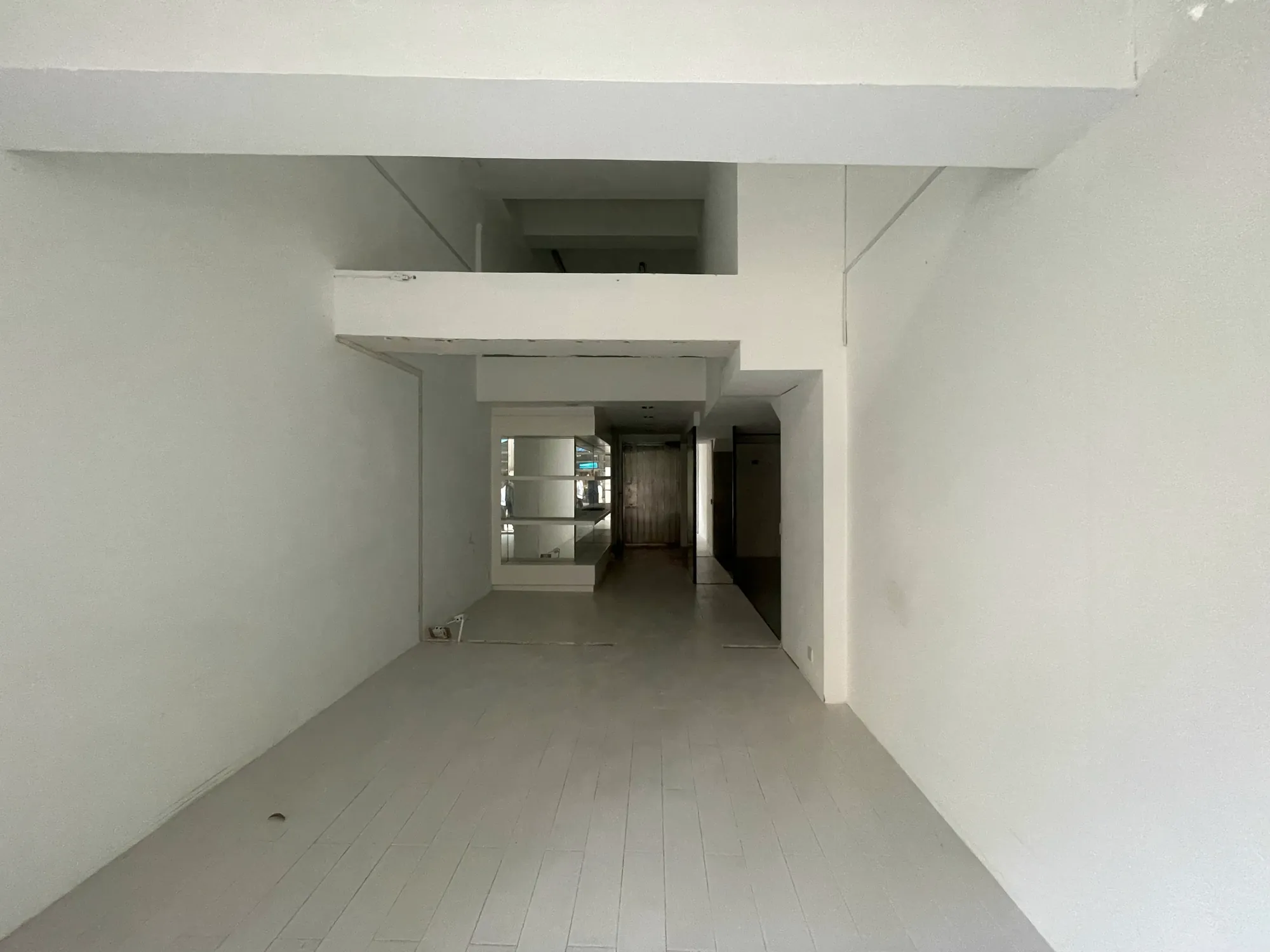 Ref#3005: A mostly empty, narrow room with white walls, ceiling, and floorideal for use as an office or shop. Features include an elevated platform, mid-room ceiling beam, back doorway, and mirrors that enhance lighting.