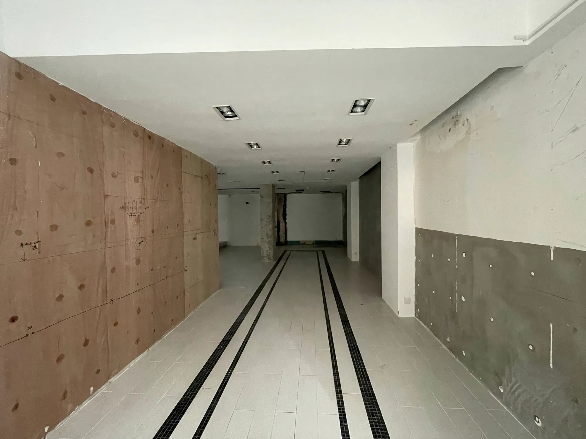 Ref#3005: A spacious, unfinished room with white tiled floors, two black floor grates, brown and gray walls, and recessed ceiling lightsideal to rent as an office or shop during construction or renovation.