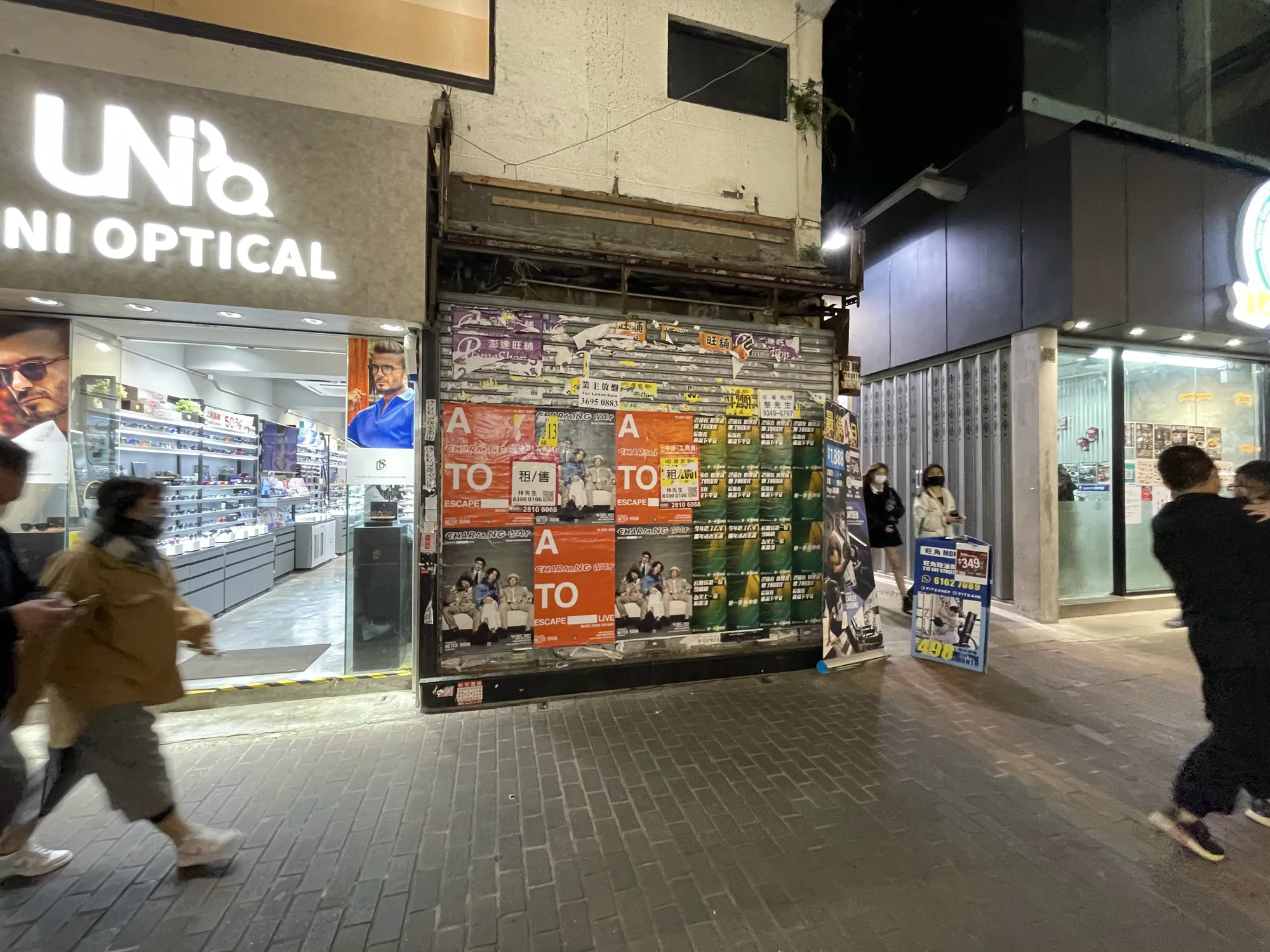 A busy street at night shows people passing a closed shop covered in colorful postersRef#3159 available for rentnext to a brightly lit optical store with glasses displays in the window.