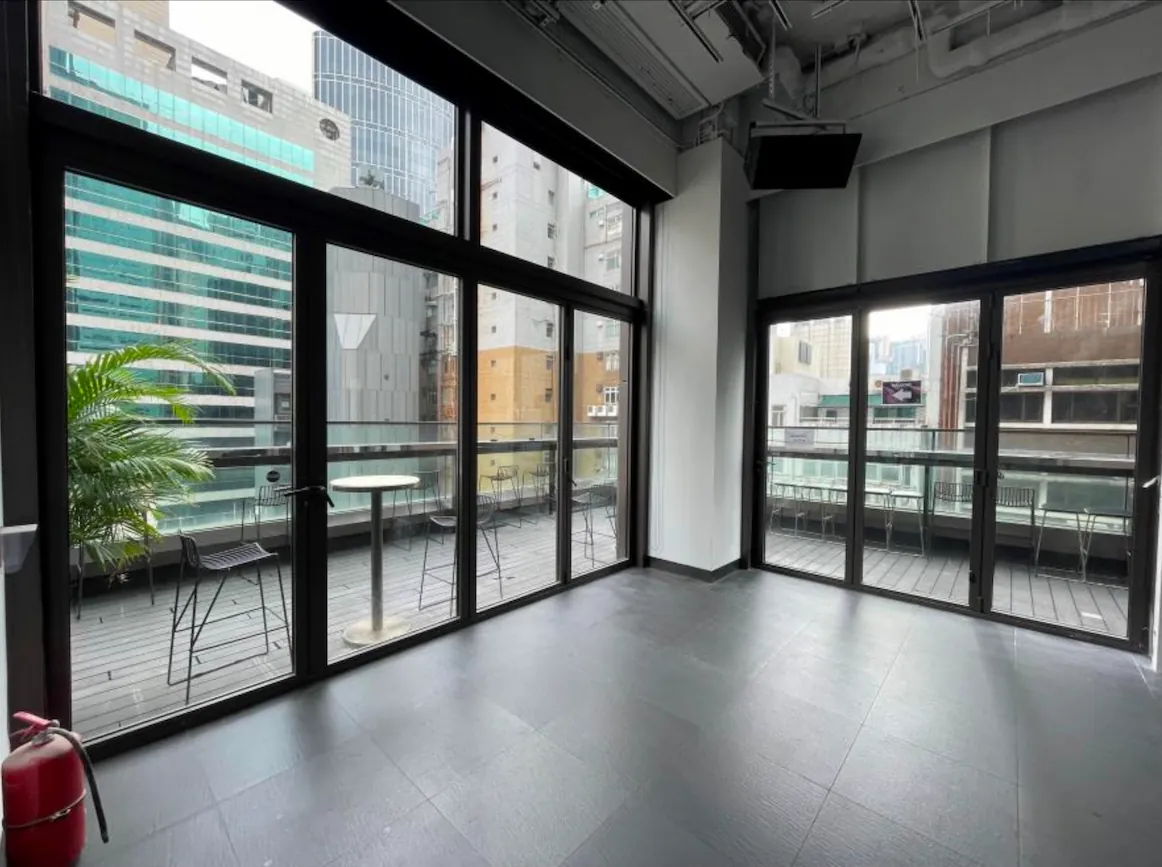 Ref#3601: Modern empty room with floor-to-ceiling windows and glass door to balconyideal for office or shop rental. Enjoy city views, outdoor tables, chairs, and a potted plant among tall buildings.