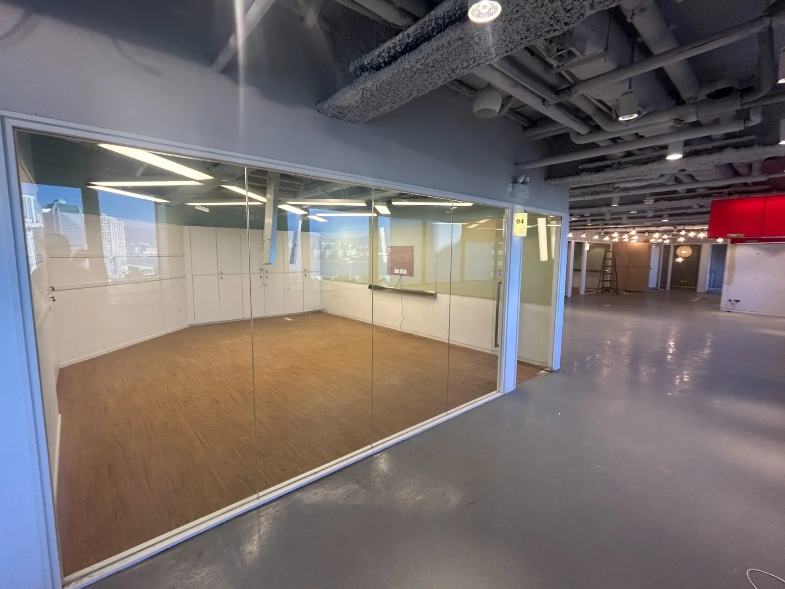 Ref#3605: Modern office space for rent with a glass-walled meeting room (wood flooring inside, gray outside), exposed pipes and lighting, an open area with red and white walls, plus string lights overhead.