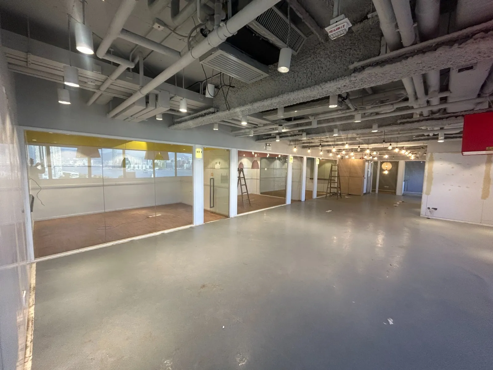 Ref#3605: Spacious unfinished office or shop space for rent, featuring exposed ceiling pipes, glass-walled rooms on one side, construction materials, ladders, mostly bare floor, and abundant natural light from large windows.