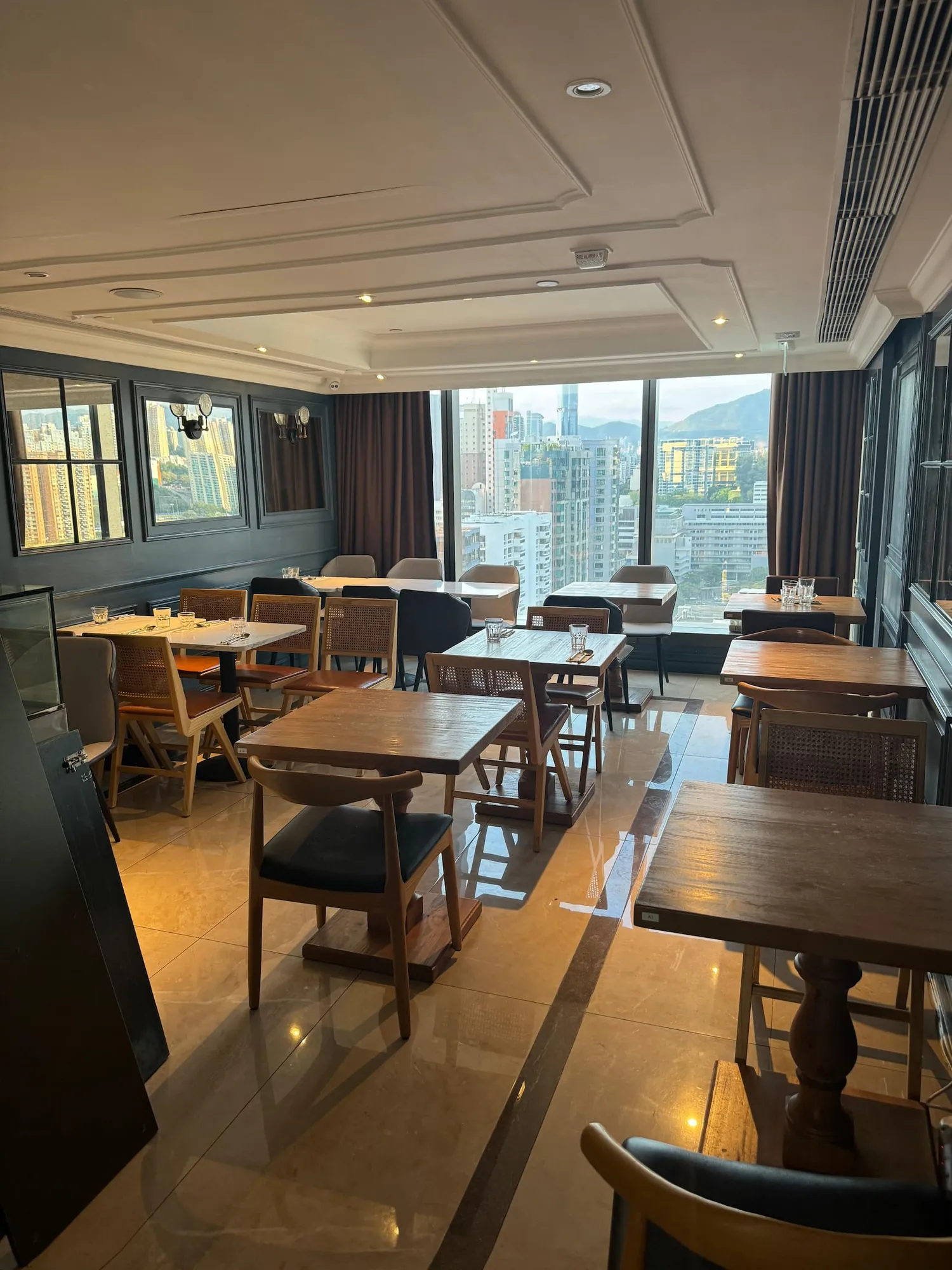 Ref#3612: A modern, empty dining area with wooden tables, chairs, large windows offering a city skyline view, and soft natural lightperfect for premium restaurant rental opportunities.