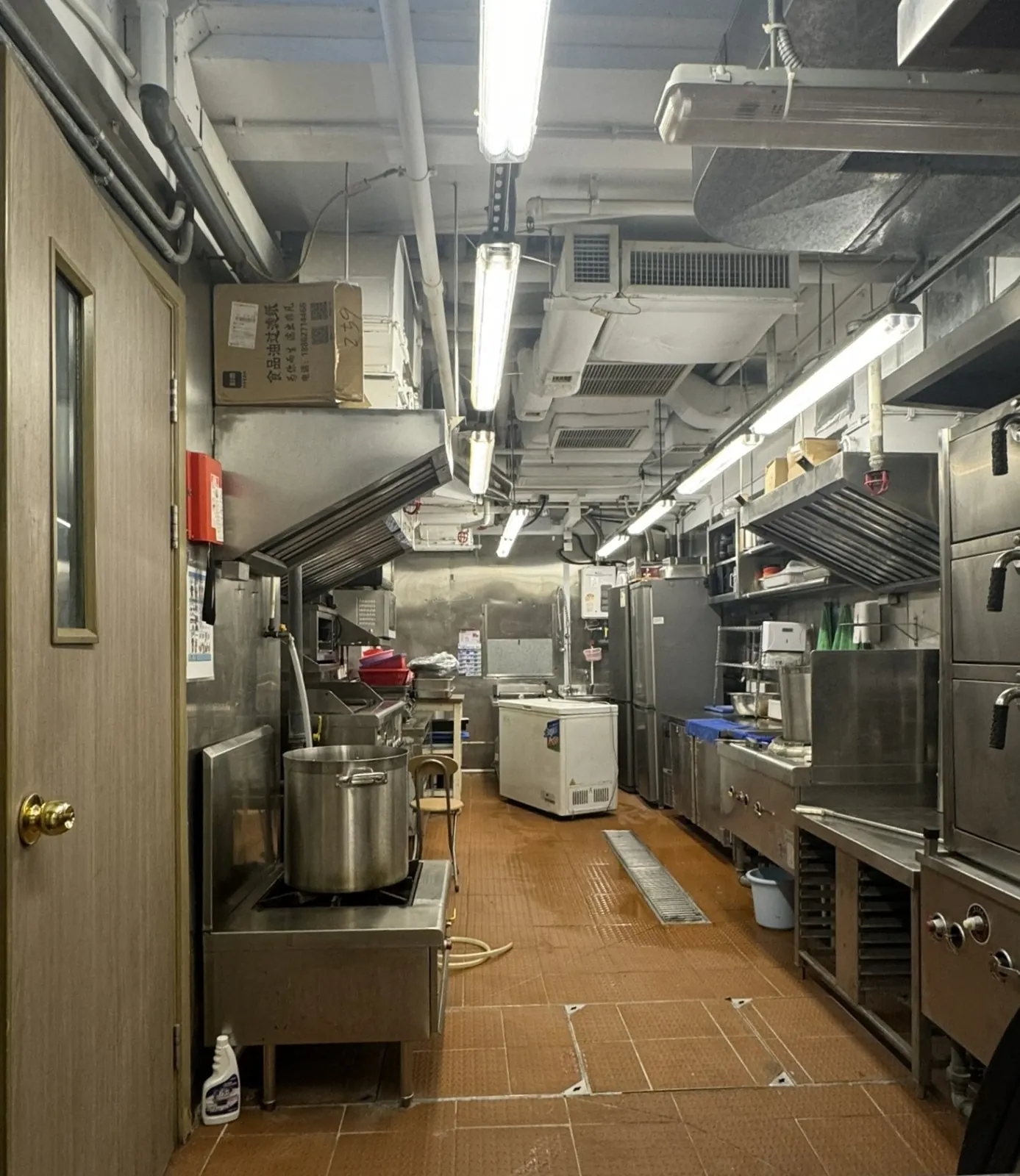 Ref#3613: Brightly lit commercial kitchen with stainless steel appliances, large pots, overhead vents, shelves, a wooden door on the left, and a tiled floor—ideal for shop rental.