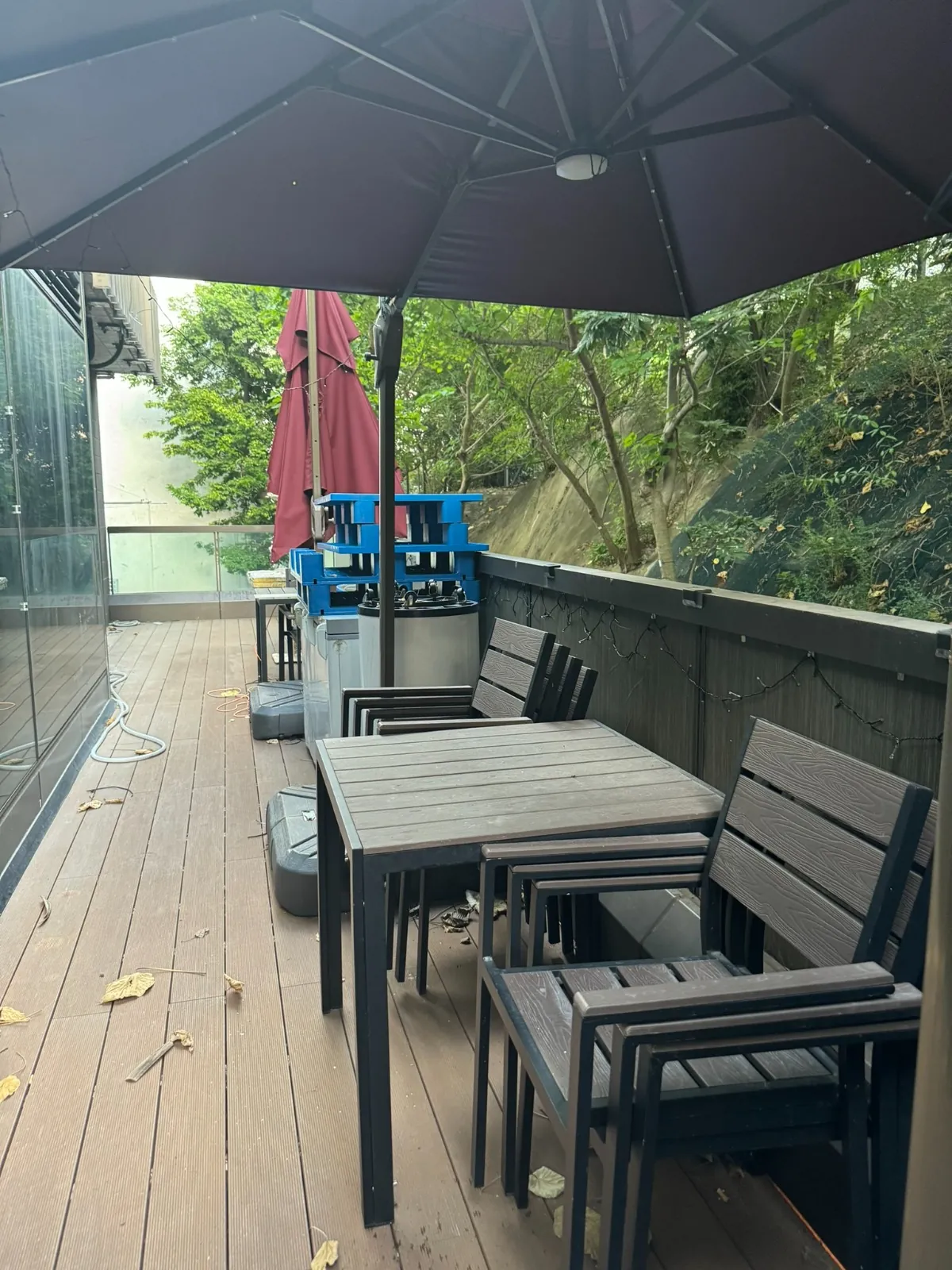 Ref#3613 features an outdoor deck with dark wooden tables, chairs, a large black umbrella, two red umbrellas, blue trays near a metal counter—all surrounded by greenery and scattered leaves on the wooden floor. Ideal for restaurant rental.