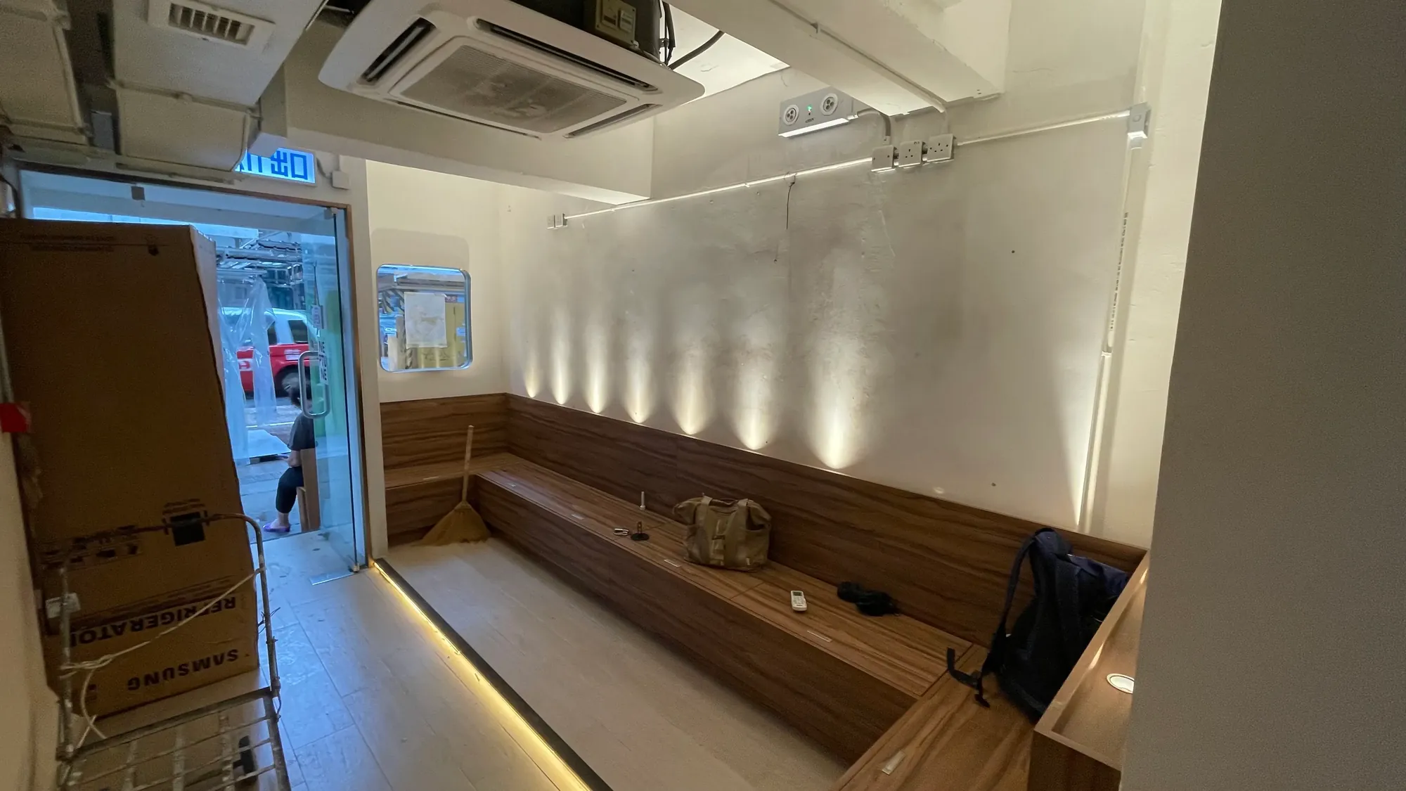 Ref#3647: Small, modern room with wood bench seating, soft wall lighting, ceiling AC, and bags on benches. Open door leads to street—perfect for use as a restaurant or shop.
