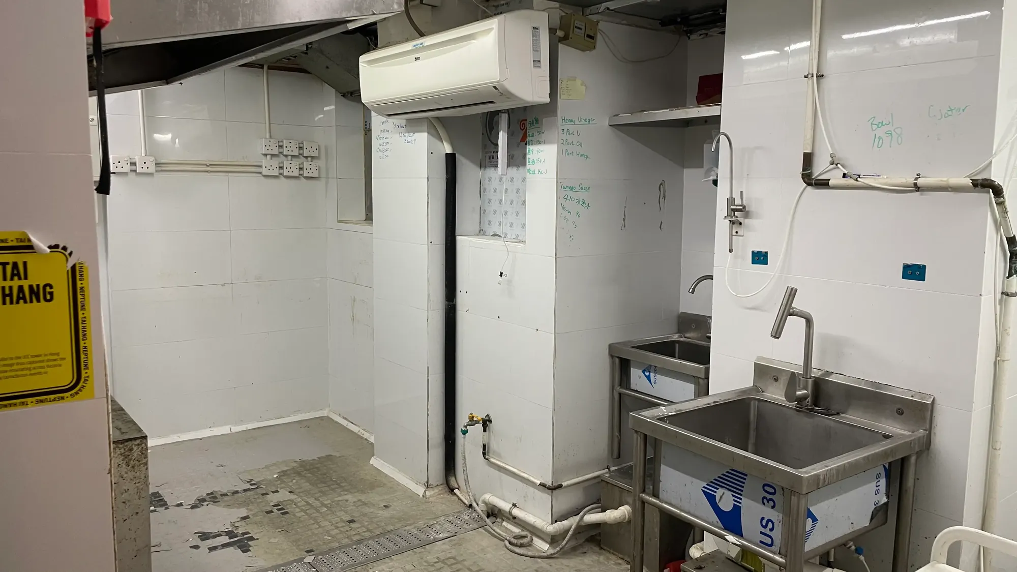 Ref#3647 is a clean industrial kitchen for rent with white tiled walls, stainless steel sinks, exposed pipes, air conditioning, and note-covered walls. A yellow caution sign is partly visible to the left. Perfect for restaurant use!.