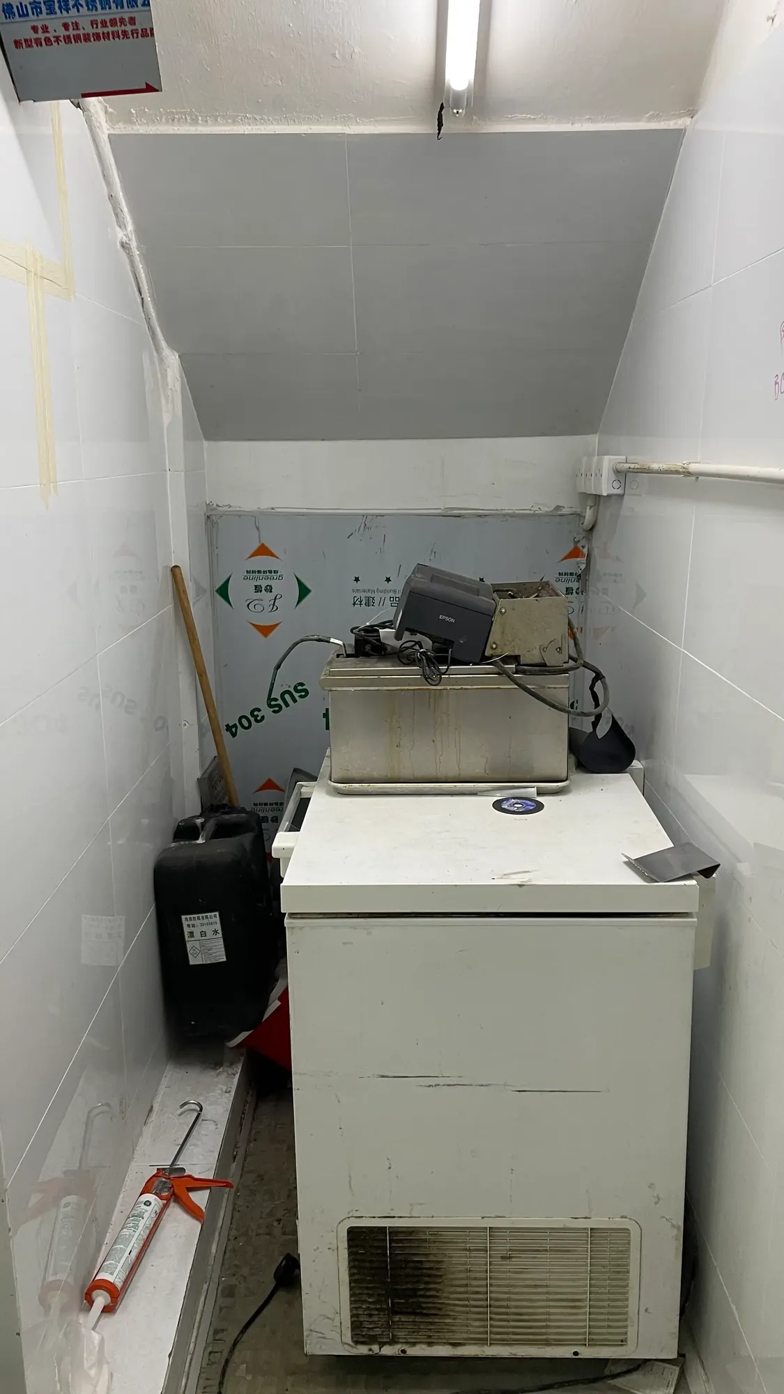 A compact storage room in a rental shop holds Ref#3647 (white chest freezer), an old cash register, cleaning products, a black bin, and is lit by a ceiling light. White walls display visible stains and markings.