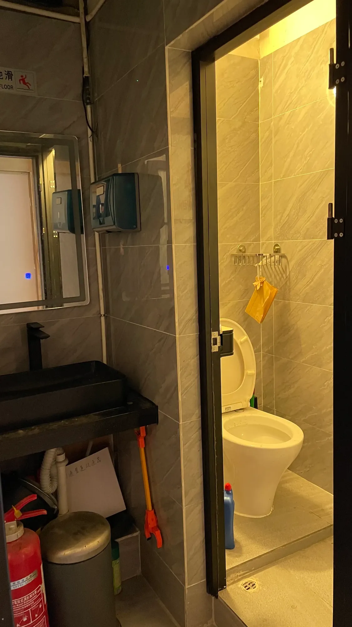 Ref#3647 features a bathroom with an open door, white toilet, yellow cleaning gloves above it, and nearby cleaning supplies—perfect for those seeking to rent offices or shops with amenities like a sink, mirror, fire extinguisher, mop, and visible pipes.