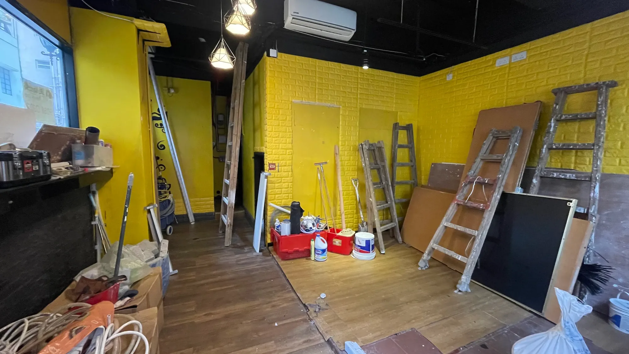 Ref#3648: Room under renovation, perfect for a restaurant or shop. Features yellow brick-pattern walls, wooden ladders, paint cans, tools, boards on wood flooring, and pendant lights on a black ceiling.
