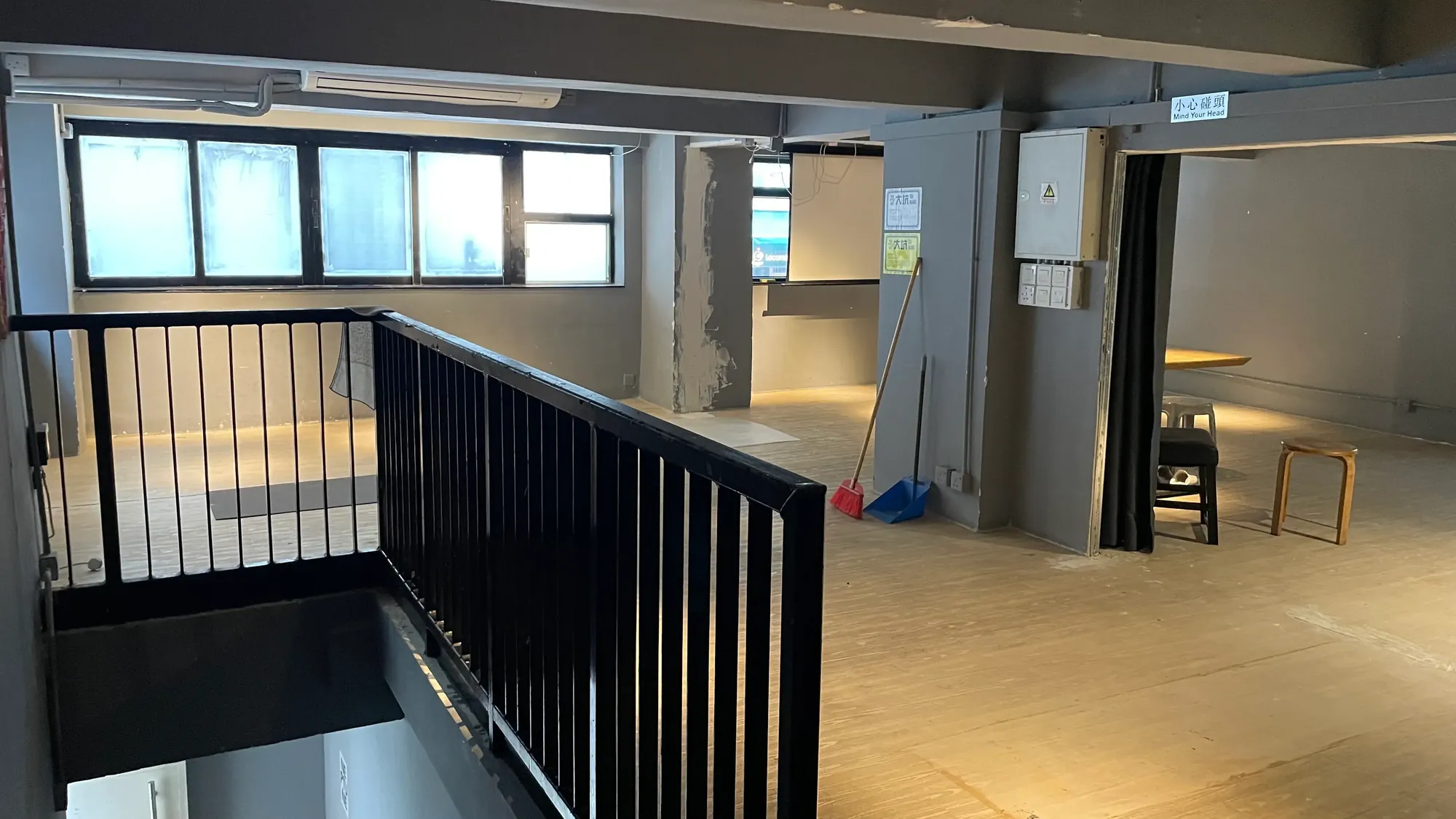 Ref#3650: Bright empty loft with large windows, gray walls, black railing, wood floors, cleaning supplies in the corner. Ideal to rent as an office or shop. Features a small table and two stools in the back.