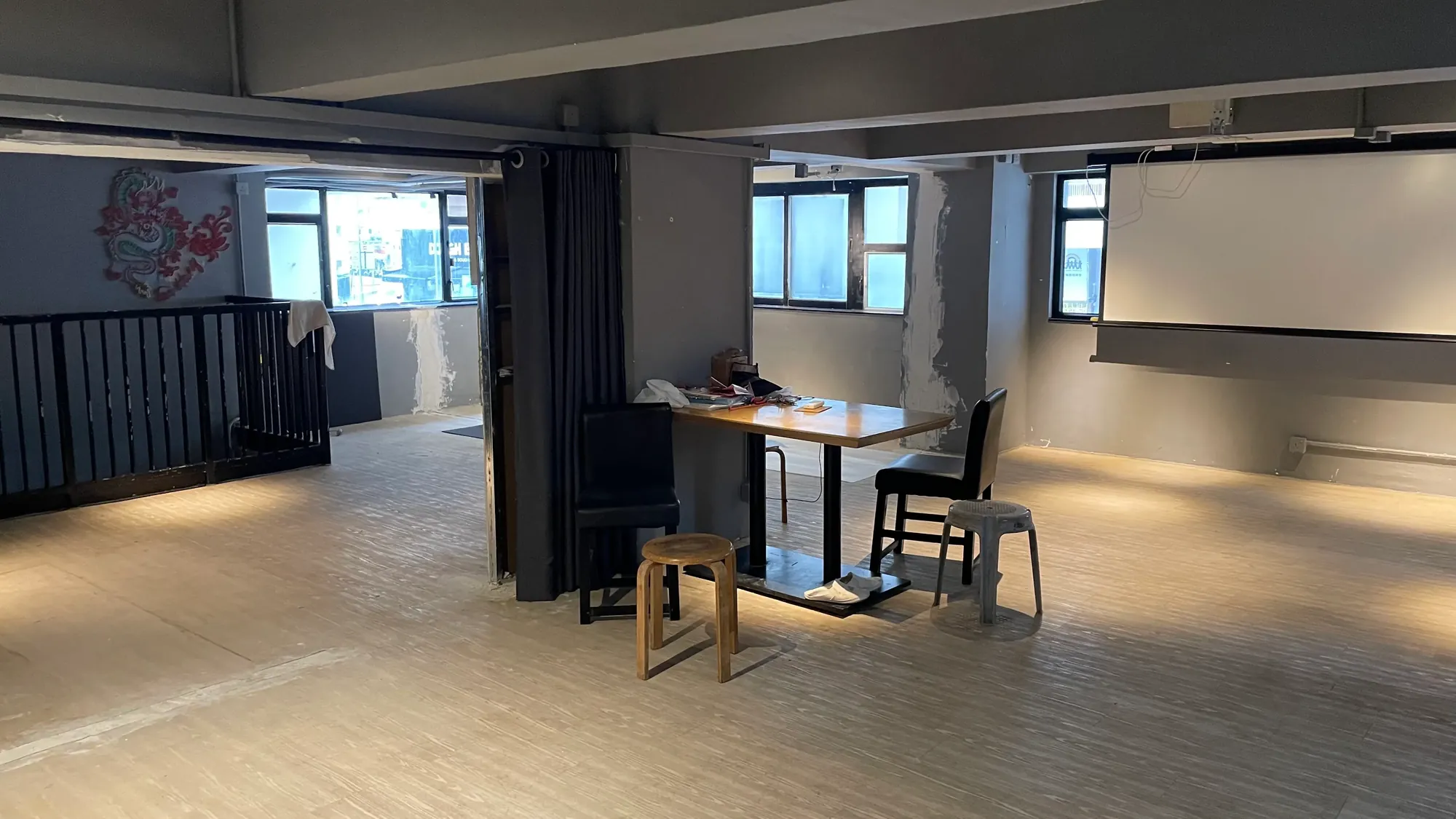 Ref#3650 is a spacious, minimally furnished room with light wood flooring, a table with two chairs, a stool, large windows with natural light, staircase railing, and a projector screenideal to rent as an office or shop.