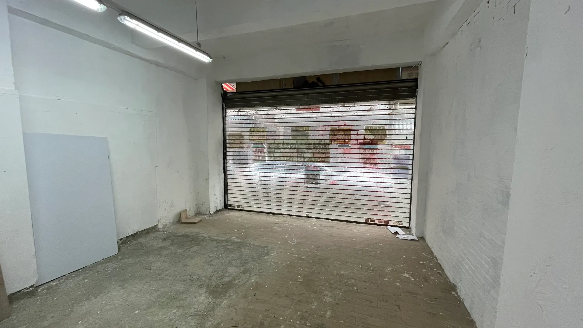 Ref#3651: Small, empty room with white walls and a slightly worn bare floor. Features a partially open metal rolling shutter door facing the street and ceiling fluorescent lightsideal for shop rental.