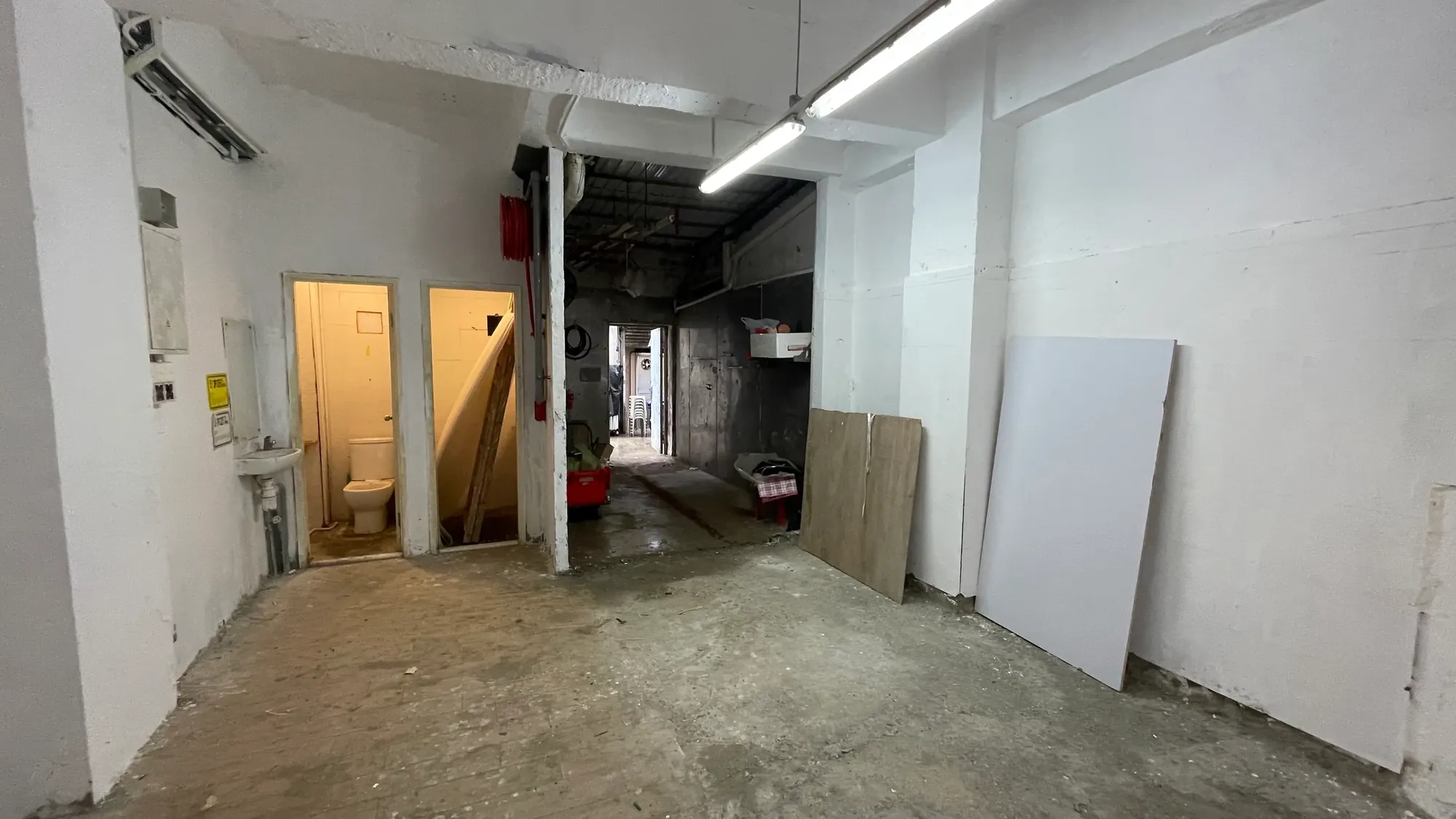 Ref#3651: Unfinished room with white walls, concrete floor, and fluorescent lightsideal for shop or office rental. Features an open bathroom door on the left and construction materials along the right wall.