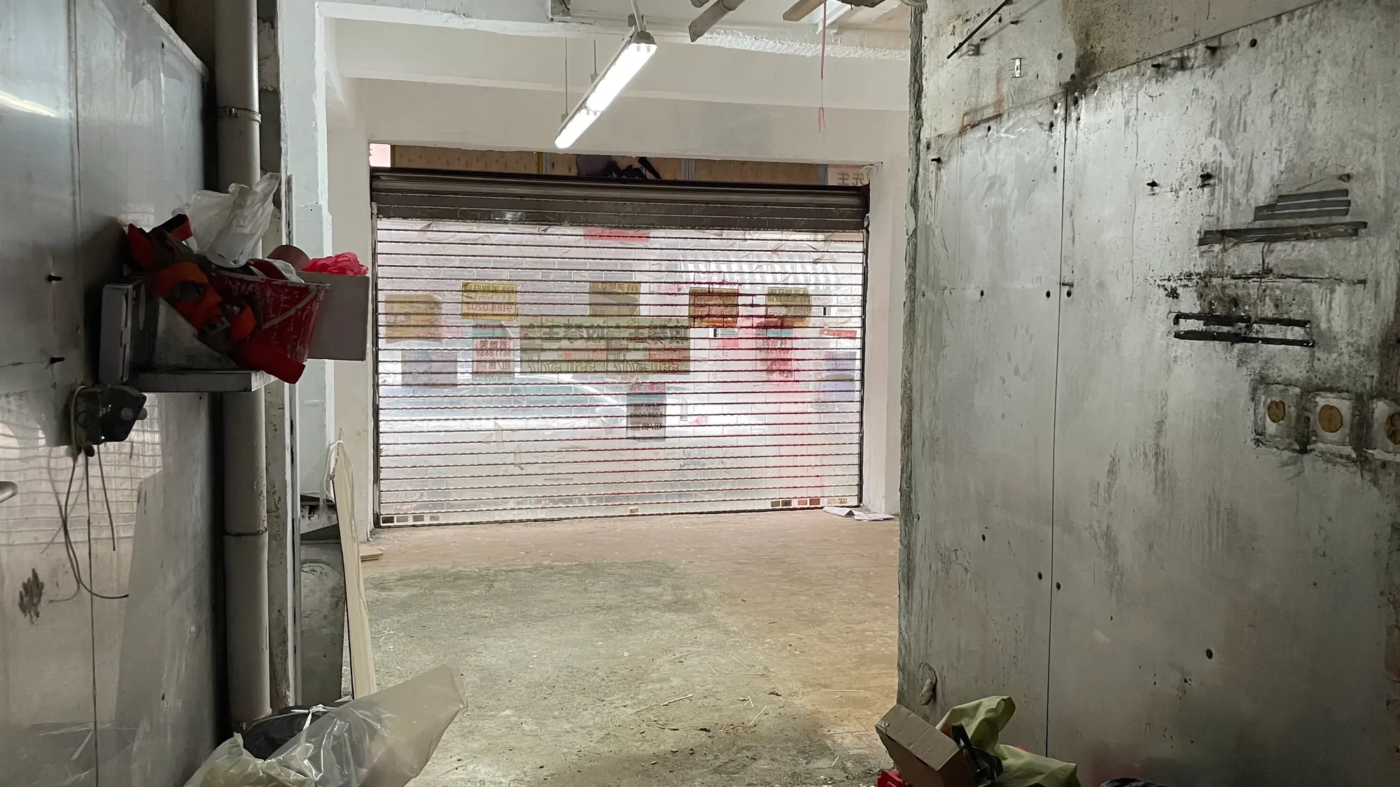 Ref#3651: An empty, unfinished garage or storage area with a partially open metal rolling shutter letting in natural lightideal for shop rentals. Various tools and objects are scattered along the walls.