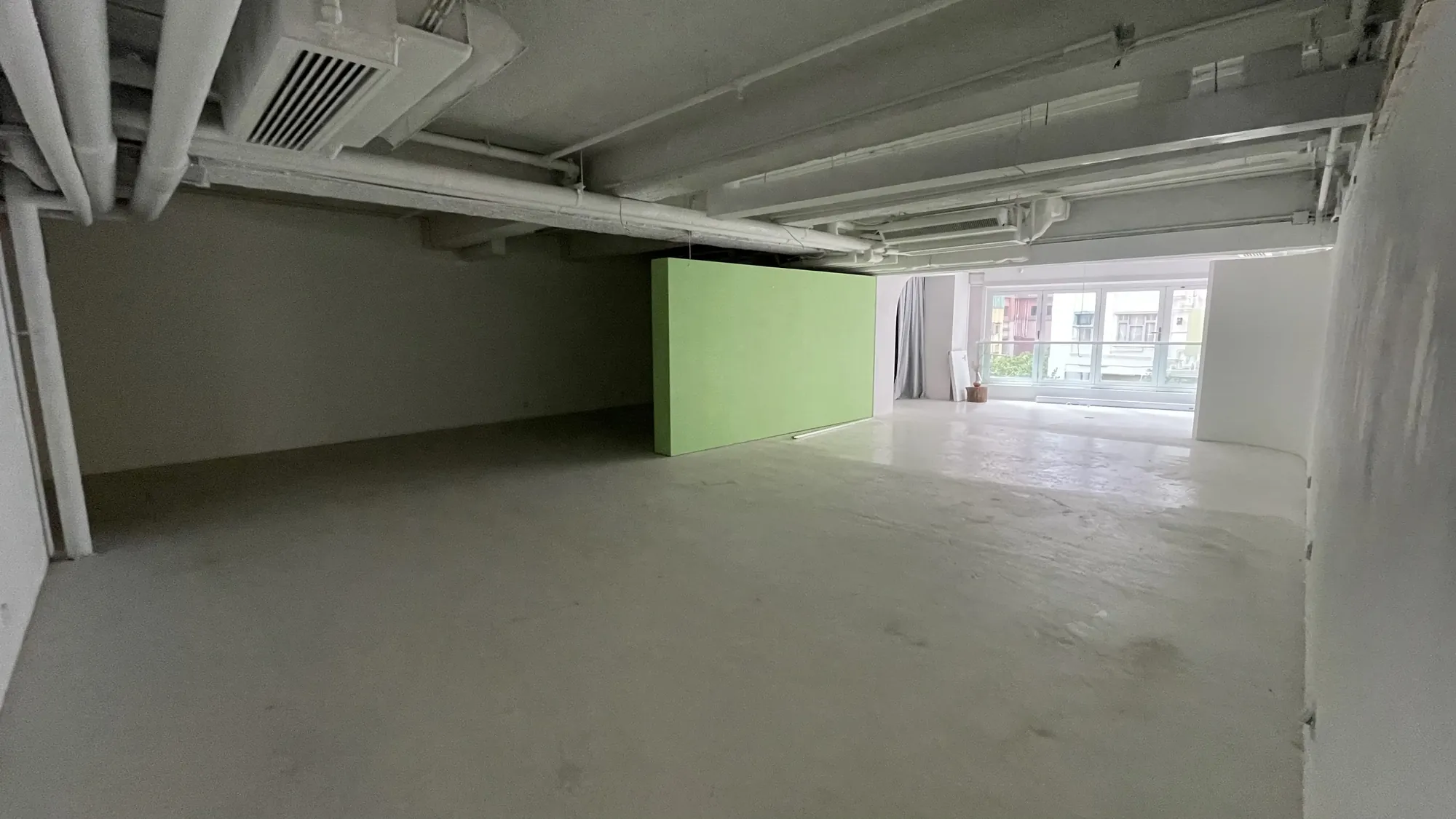 Ref#3676: A spacious, empty room with white floors and ceiling, exposed pipes, a large green divider wall, and a window bringing in natural lightperfect for office rental or creative uses.