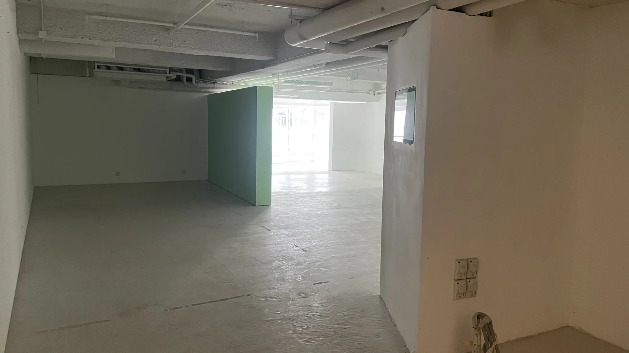 Ref#3676: Spacious empty room with white walls and floors, exposed ceiling pipes, green central partition, and large windows for natural lightideal to rent as a shop or office space.