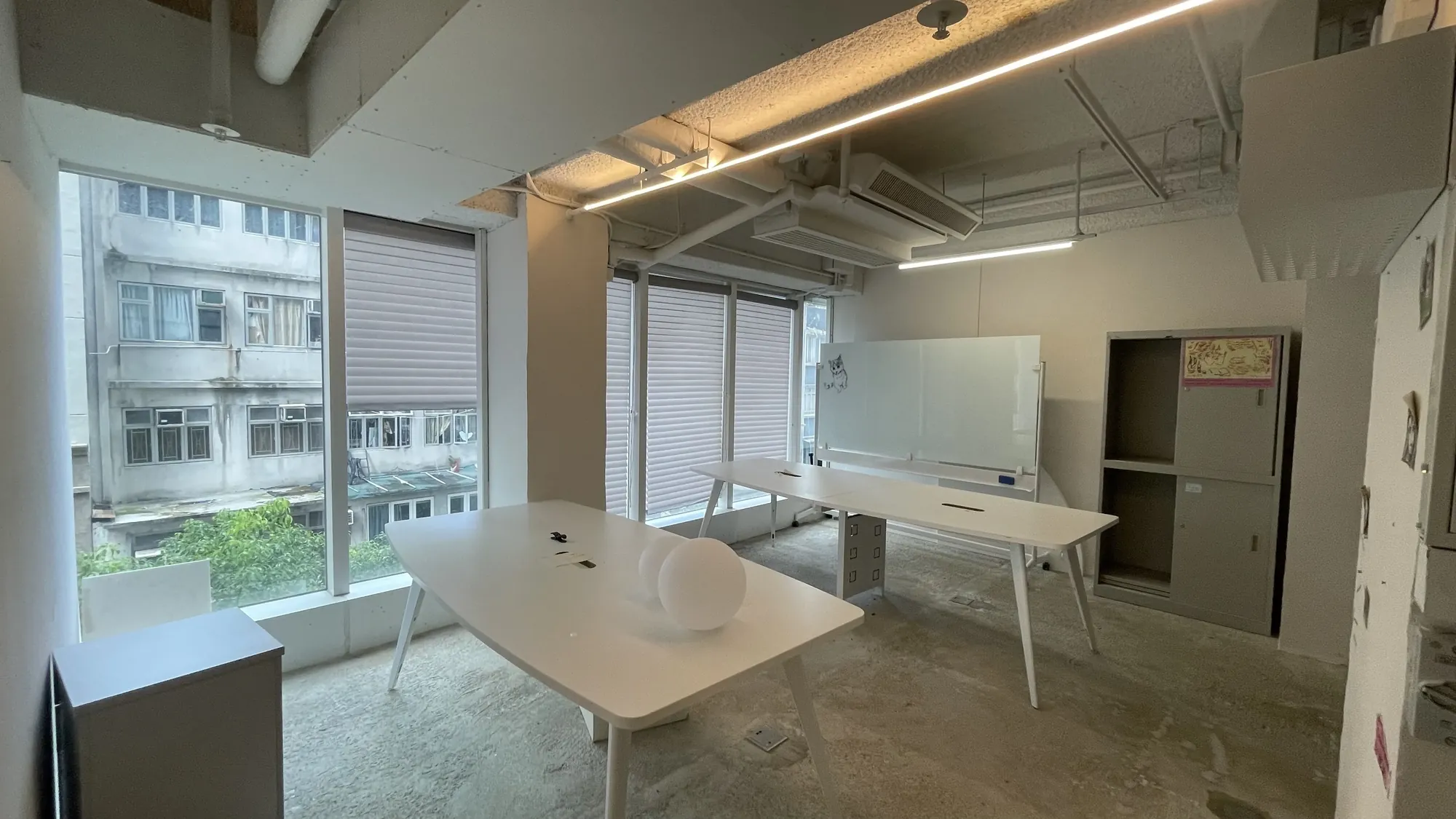 Ref#3676: Modern minimalist office for rent with large windows, two white desks, whiteboard, gray cabinet, exposed ceilings, and building views. Bright, uncluttered spaceideal for your next office rental.