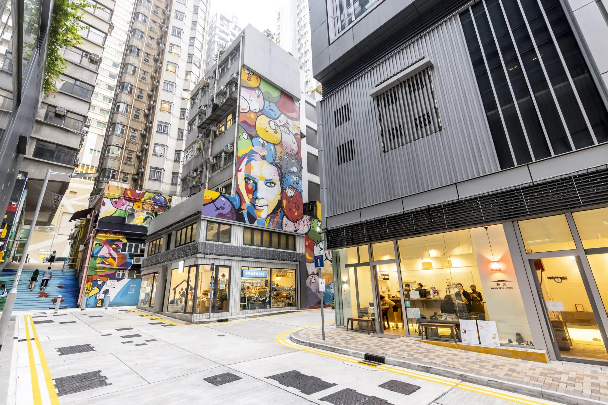 西營盤餐廳舖位。一條街道，設有色彩繽紛的壁畫、高聳大廈，以及一間有顧客在內的現代化咖啡店。