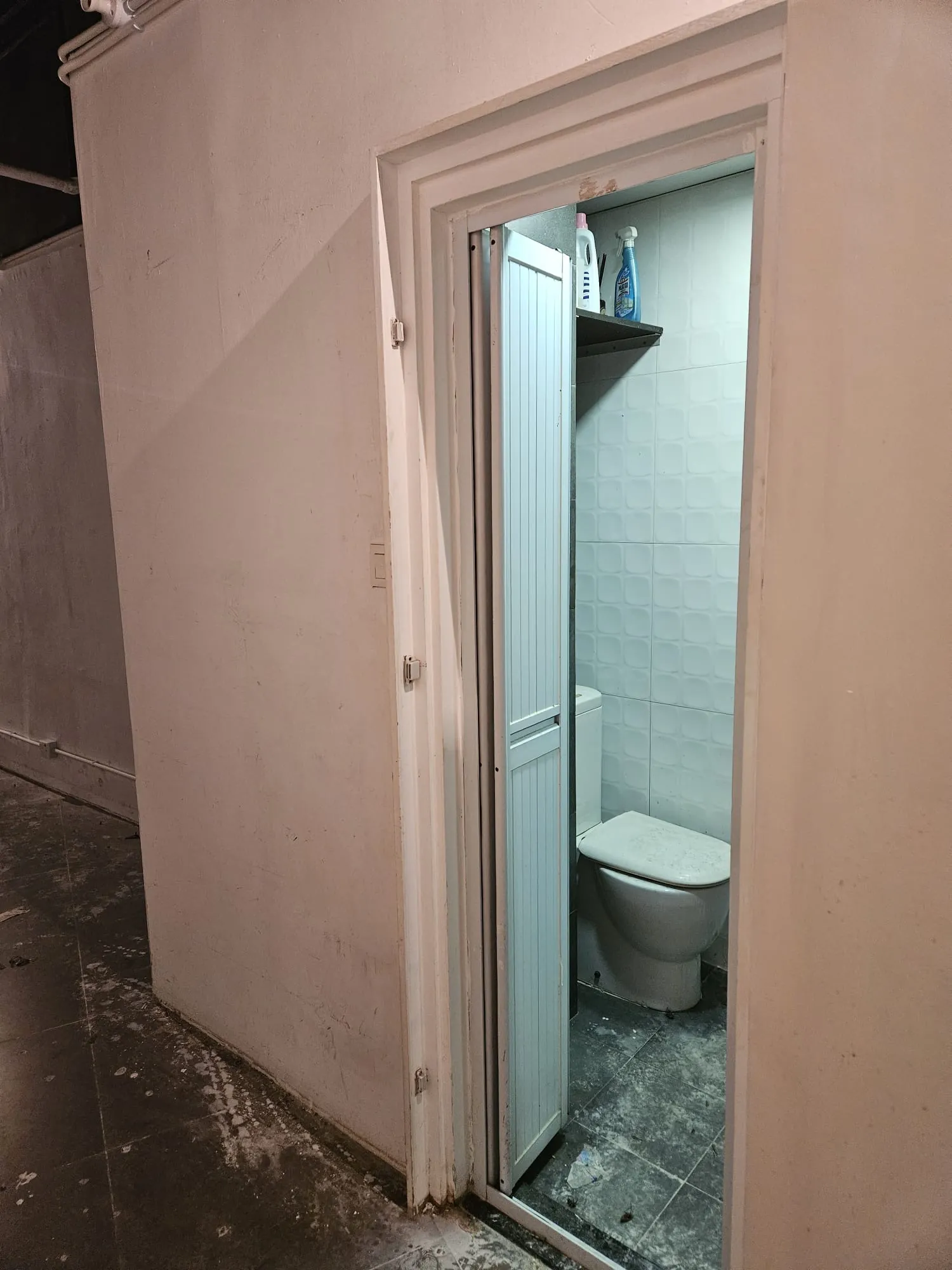 Small bathroom on Lyndhurst Terrace Central with white toilet folding door tiled walls and cleaning supplies above.