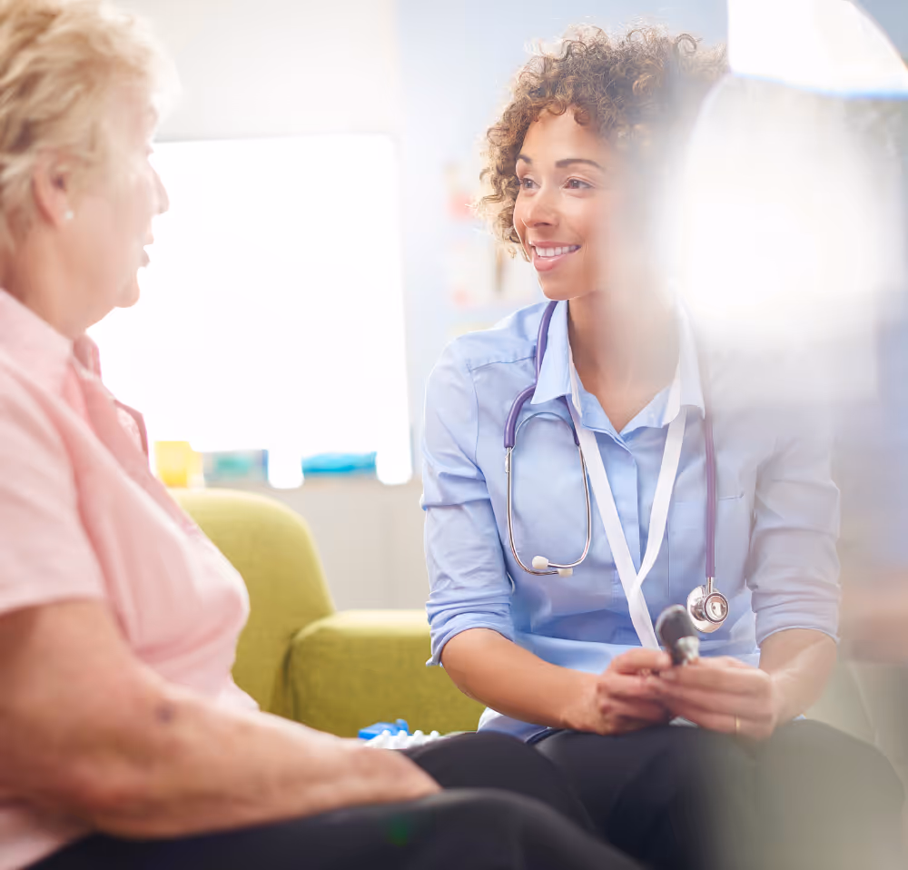 Smiling female doctor with a stethoscope speaking compassionately with an older female patient in a bright, welcoming clinic setting, highlighting patient-centred care.