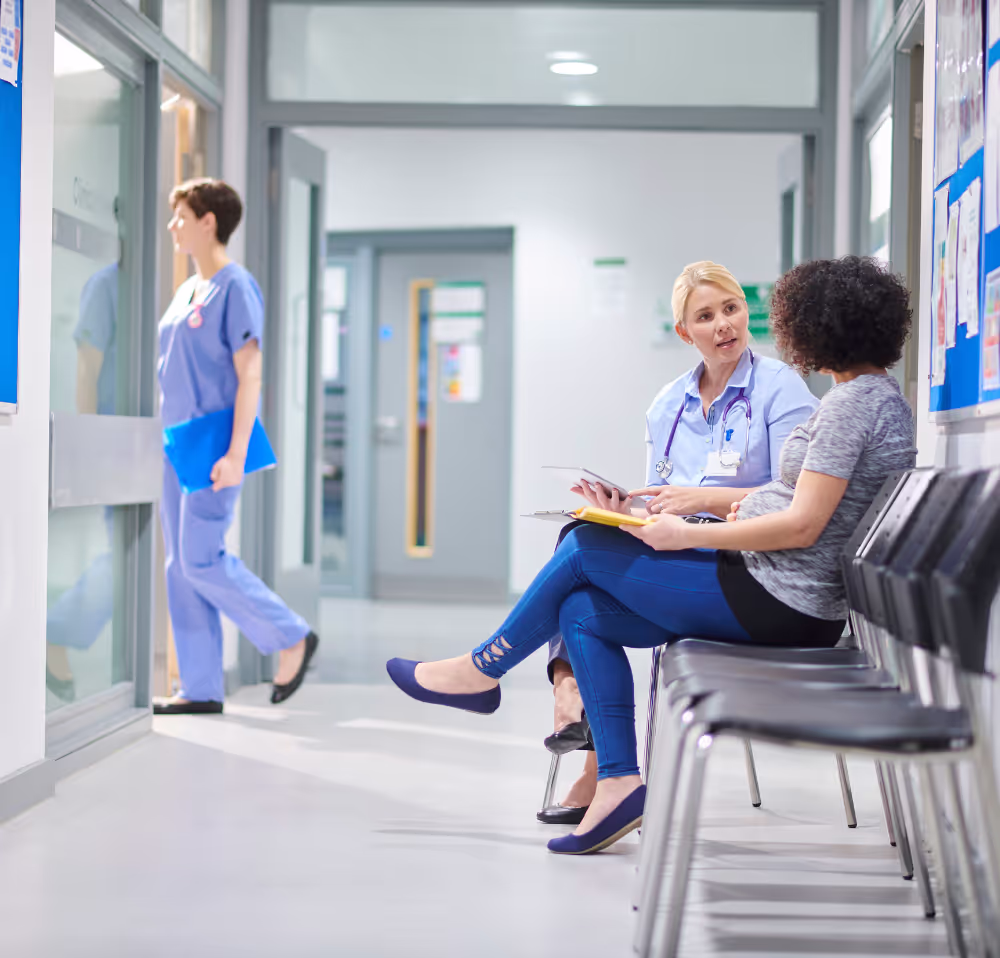 Female healthcare professional discussing medical notes with a patient in a hospital waiting area, while another nurse walks past clinic rooms in a clean, modern facility.