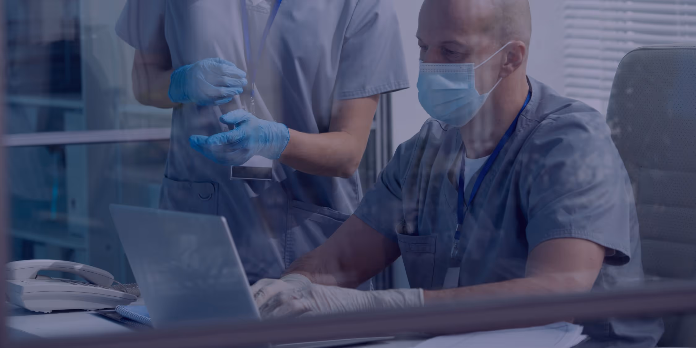 Two healthcare professionals wearing scrubs and masks working together at a desk, with one using a laptop and the other standing beside them.