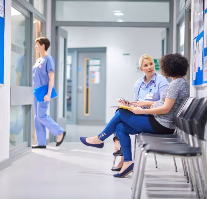 Female healthcare professional discussing medical notes with a patient in a hospital waiting area, while another nurse walks past clinic rooms in a clean, modern facility.