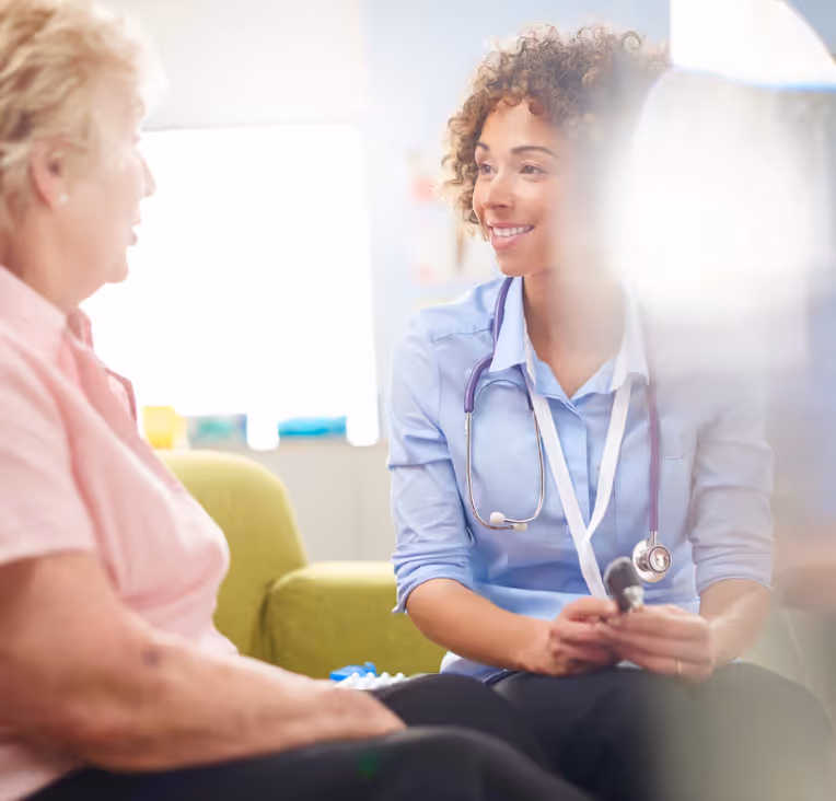 Smiling female doctor with a stethoscope speaking compassionately with an older female patient in a bright, welcoming clinic setting, highlighting patient-centred care.