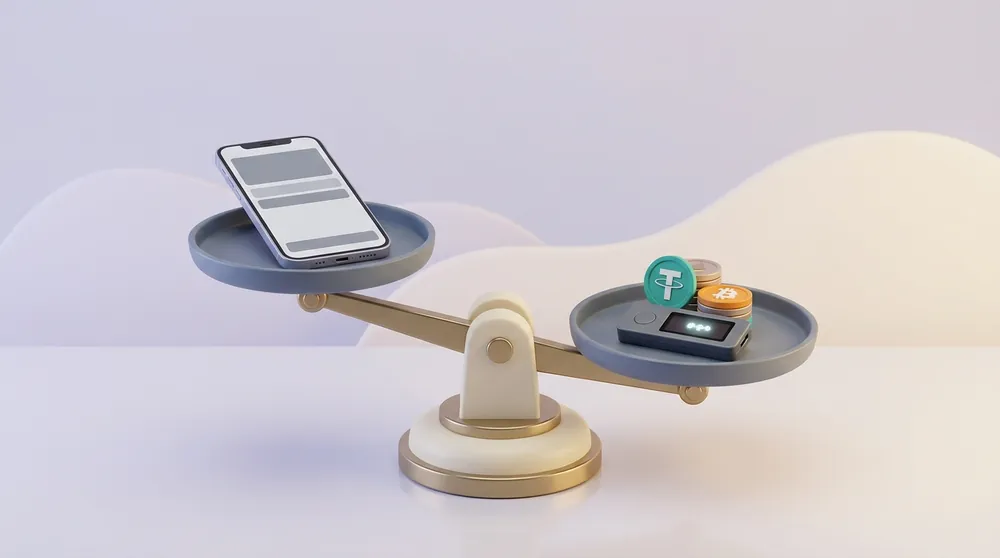 A balance scale on a desk: a smartphone showing a wallet screen sits on the higher pan, while a small hardware device under a stack of bitcoin and Tether coins weighs the lower one down