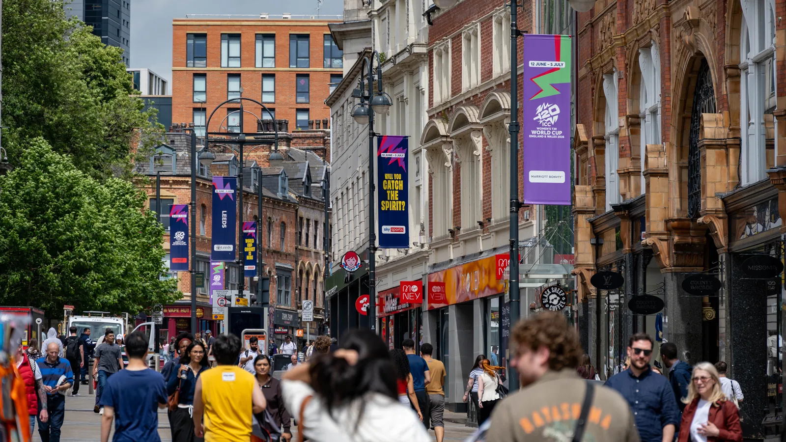 Purple and pink ICC Women's T20 World Cup lamppost banners lining Briggate in Leeds above a busy shopping street.
