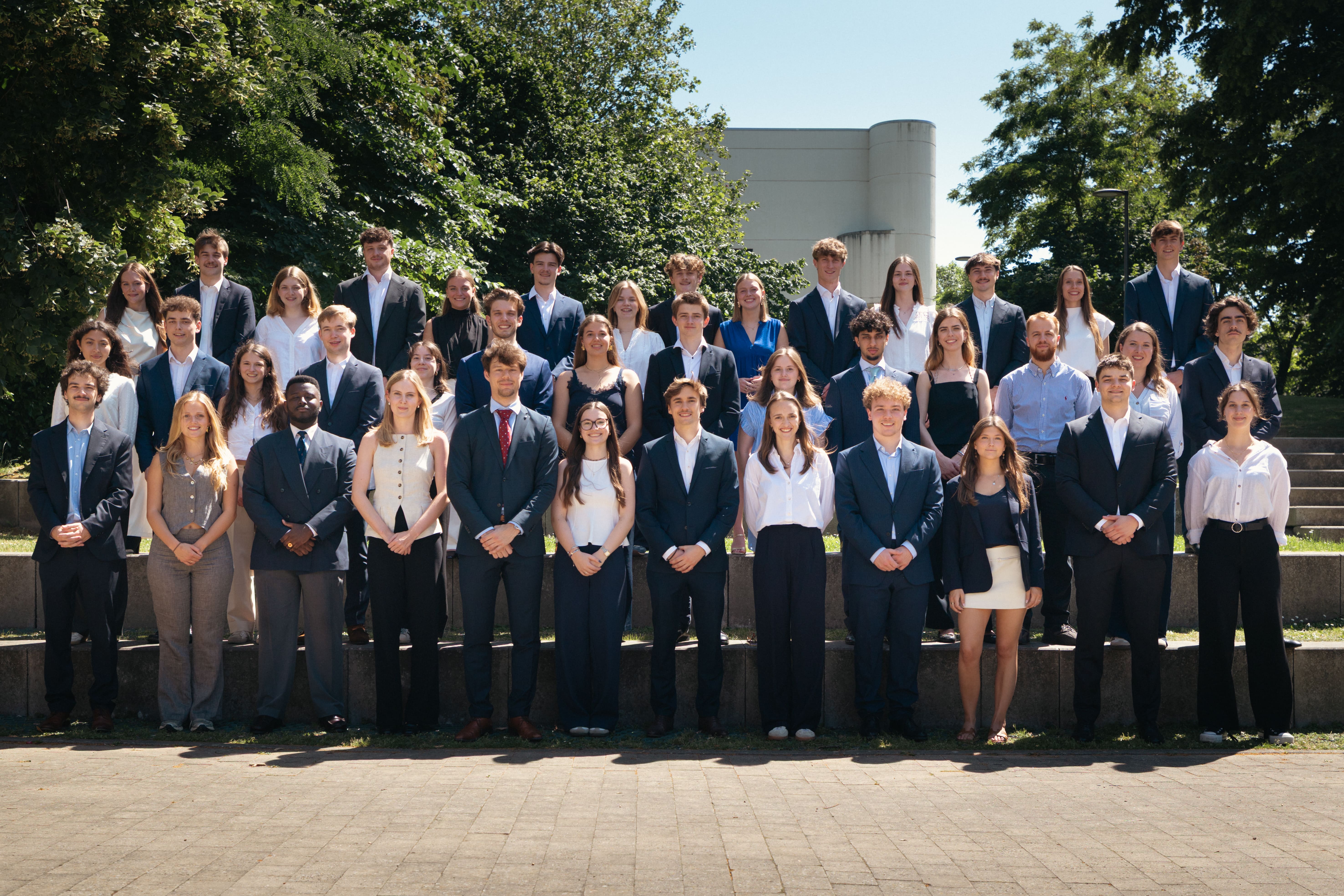 Group of around 45 young adults dressed in formal attire posing outdoors on concrete steps with trees in the background, many showing hand signs and smiling.