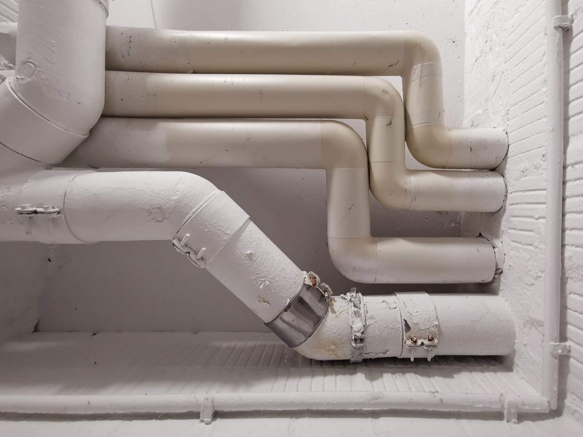 White painted interconnected pipes with joints mounted against a white brick wall.