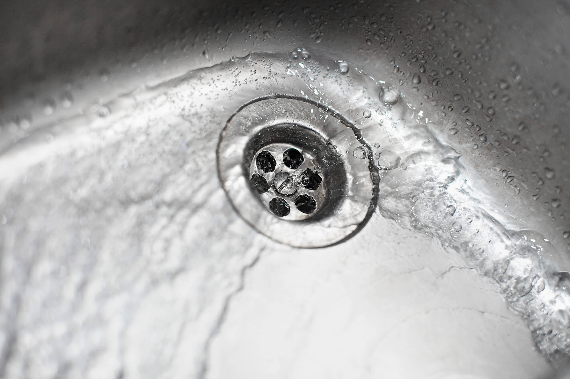 Close-up of water flowing into a stainless steel sink drain with water droplets on the surface.