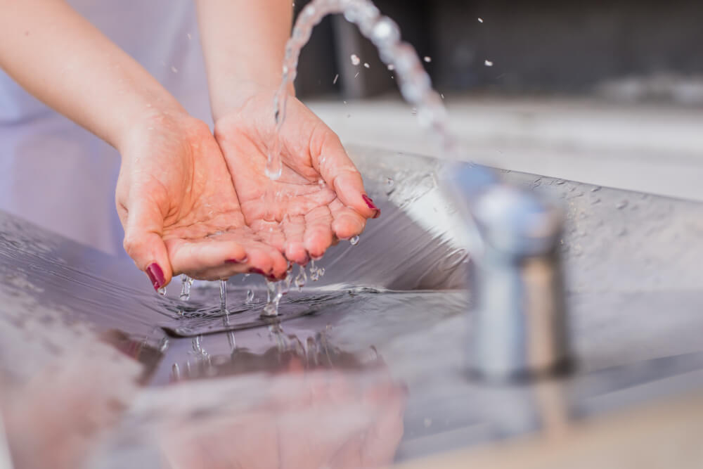 Close-up of hands cupping running water from a metal faucet over a stainless steel sink.