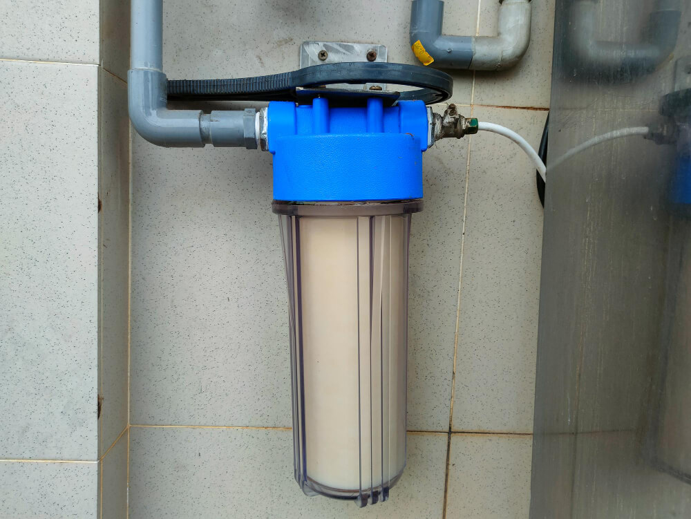 Blue water filter housing mounted on a tiled wall with connected grey pipes and a black circular handle on top.